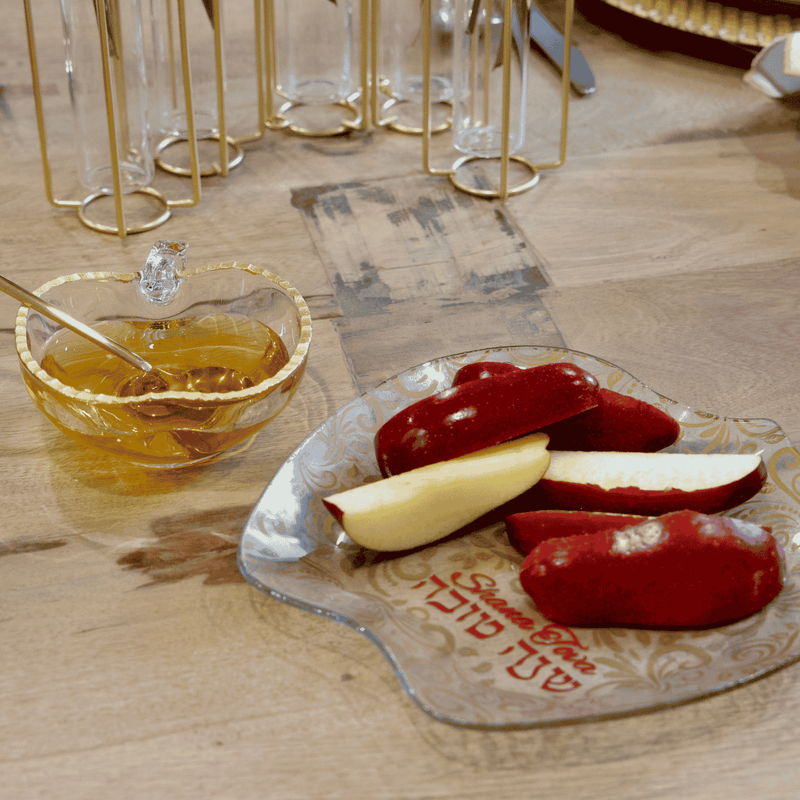 A styled gourmet gift basket or charcuterie board arrangement on a wooden table, featuring a black tray with assorted nuts, dried fruits, crackers, a bottle of olive oil, and packaged sweets, alongside branded napkins and serving dishes.