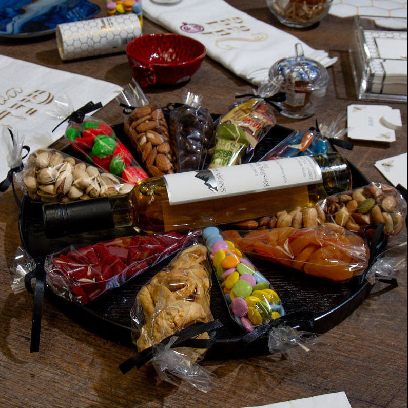 A styled gourmet gift basket or charcuterie board arrangement on a wooden table, featuring a black tray with assorted nuts, dried fruits, crackers, a bottle of olive oil, and packaged sweets, alongside branded napkins and serving dishes.
