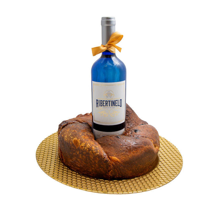 A bottle of Ribertinelo white wine with a blue glass body, silver foil, and a yellow ribbon bow, placed upright on top of a large round brown babka on a gold textured circular plate, against a solid white background.
