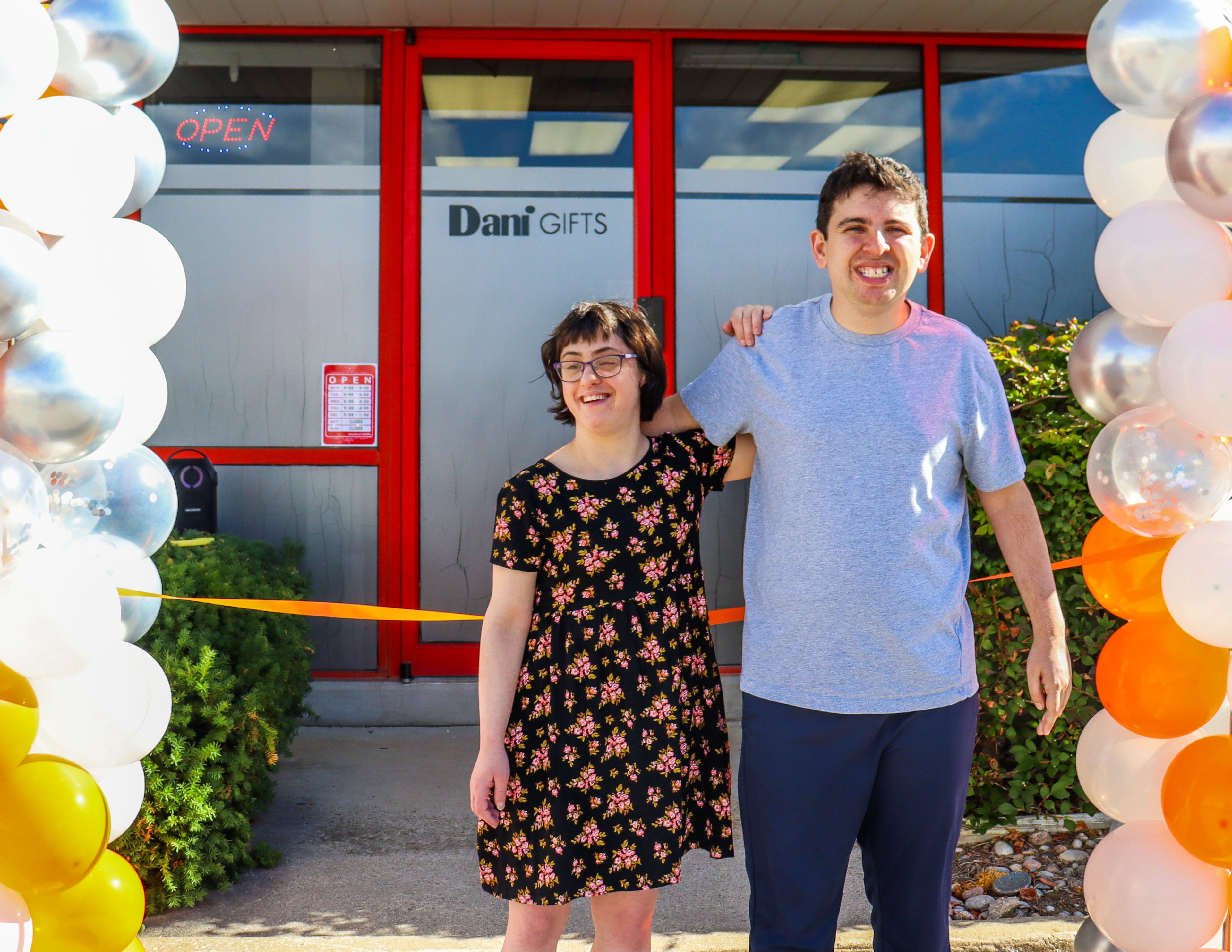 Two people standing in front of a store with balloons on either side.