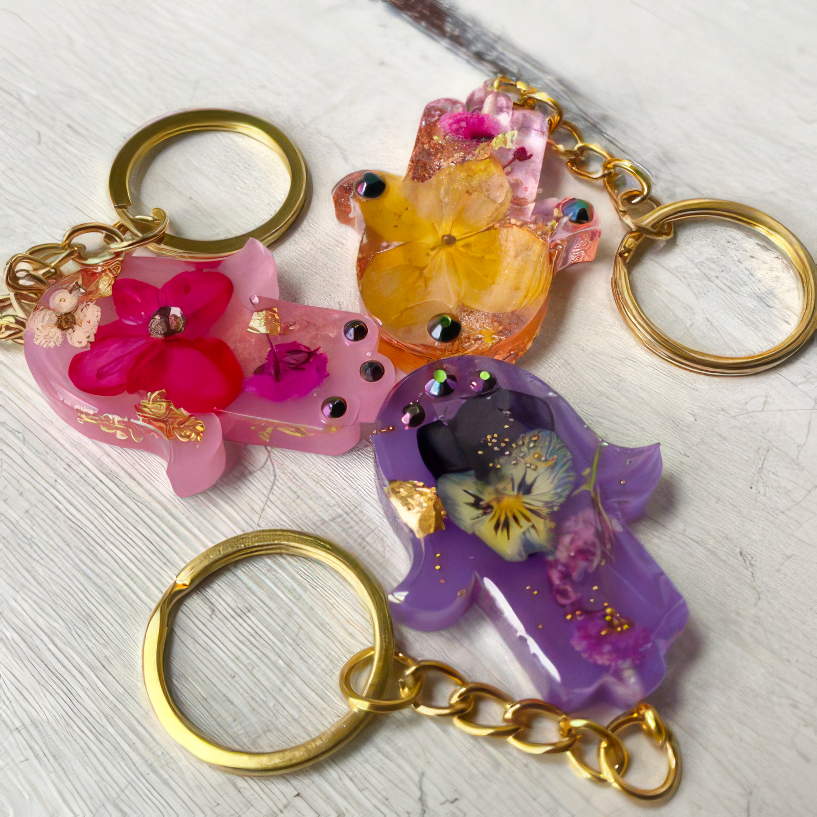 Three colorful hamsa hand keychains with gold-tone chains and rings, each containing a dried flower and gold leaf, displayed on a light wood background.