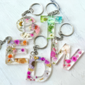 A set of letter-shaped keychains arranged on a white textured surface. Each keychain features a clear resin letter filled with pressed flowers and small gold leaf accents, attached to a silver metal key ring.