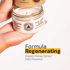 A hand holds an open jar of beeswax candle labeled 'Royal Cream Ultra Nourrissant' and 'Miel Regenerant' against a white background, with text below reading 'Formula Regenerating' and 'Organic Honey Extract from Provence'.