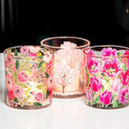 Three clear glass candle holders with pink floral patterns, including roses and tulips, arranged on a white surface against a dark background.