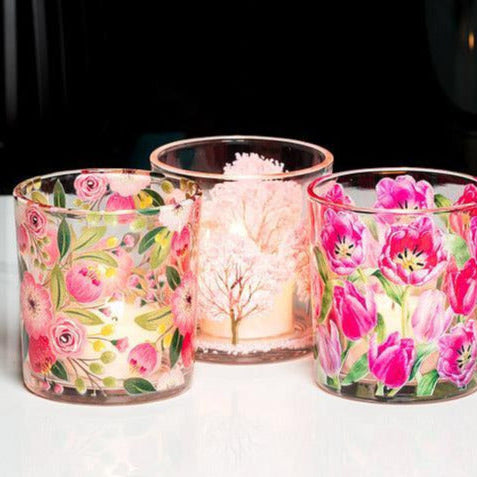 Three clear glass candle holders with pink floral patterns, including roses and tulips, arranged on a white surface against a dark background.