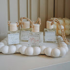 Three small clear glass perfume bottles with striped white and beige caps and matching reed diffusers on a white beaded tray, set against a beige paneled background.