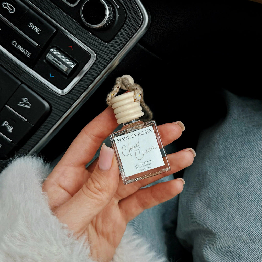 A hand holding a small glass bottle of car fragrance with a beige reed diffuser stopper, positioned near a car's center console and climate control buttons.