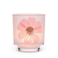 A pink glass jar candle with a large pressed pink flower embedded in the front, set against a plain white background.
