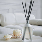 A reed diffuser bottle with black reeds sits on a light wooden table in a bright, modern living room setting with a white sofa and a floor lamp in the background.