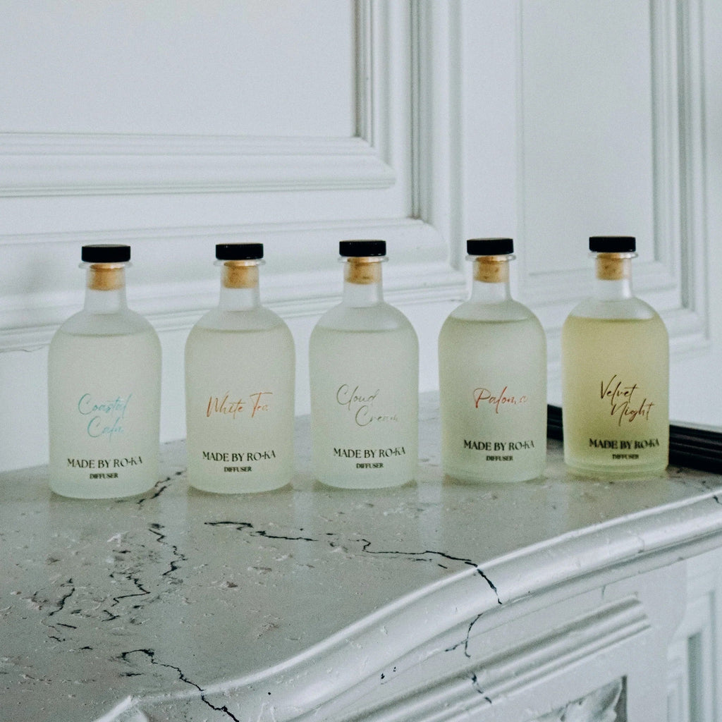 Five frosted glass perfume bottles with black caps and gold neck rings lined up on a white marble mantelpiece against a white paneled wall. The bottles contain clear or pale yellow liquids and are labeled with elegant script names including 'Cruelty Free', 'Velvet Touch', 'Scent of Roma', 'Made by Roma', and 'Pleasant'.