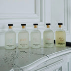 Five frosted glass perfume bottles with black caps and gold neck rings lined up on a white marble mantelpiece against a white paneled wall. The bottles contain clear or pale yellow liquids and are labeled with elegant script names including 'Cruelty Free', 'Velvet Touch', 'Scent of Roma', 'Made by Roma', and 'Pleasant'.