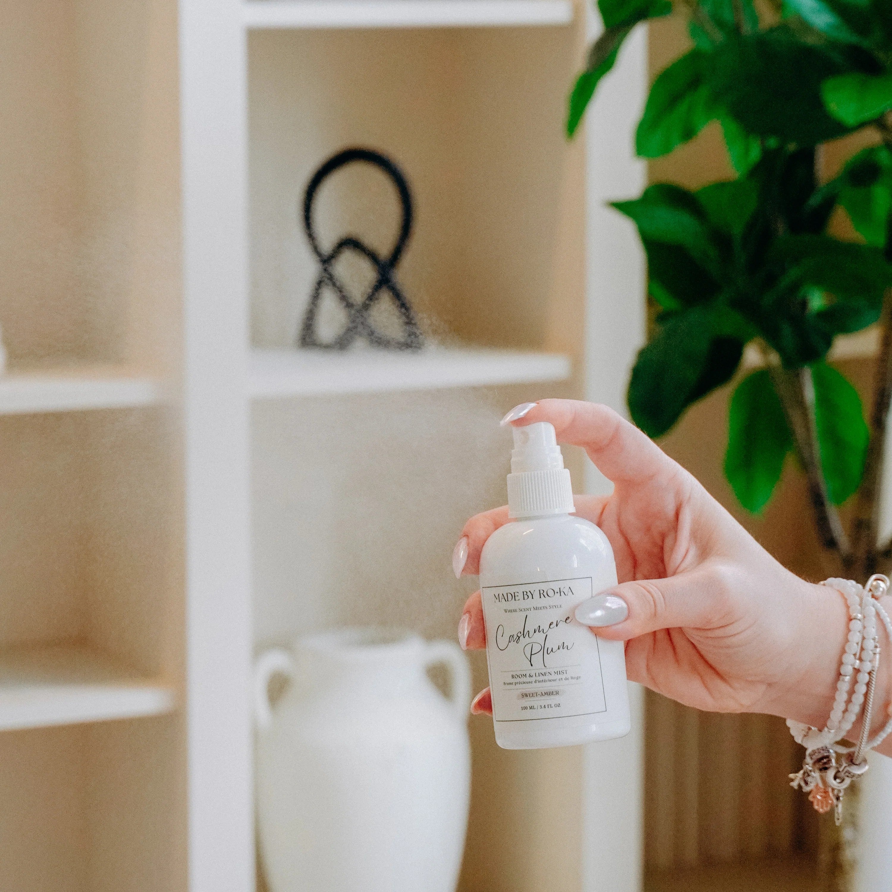 A hand holding a white spray bottle labeled 'Lavender & Mint' in a cozy, softly lit room with a white shelving unit and a green plant in the background.