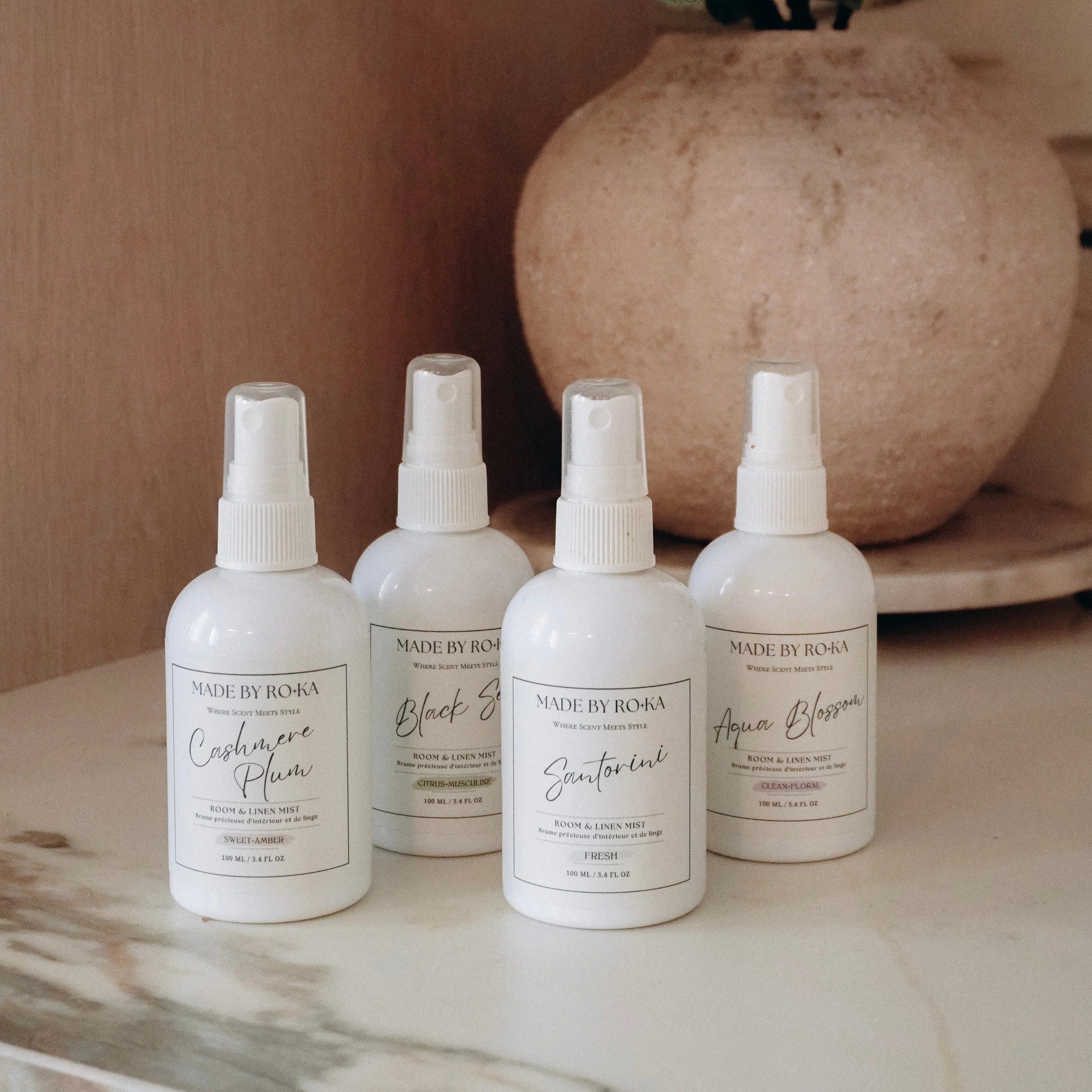 Four white spray bottles from Made by Borka Home Fragrance, arranged on a marble surface with a warm wood background and a decorative vase.