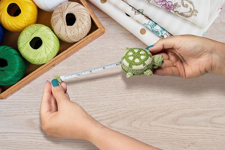 Person measuring with a small green crocheted turtle measuring tape and colorful yarn and fabric in the background.