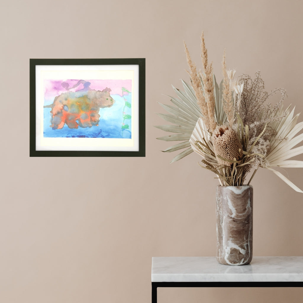 Framed artwork of a colorful bear on a beige wall with a vase of dried plants on a table.