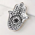 A Hamsa hand-shaped decorative tray with black floral and geometric henna-style patterns on a white background, finished with a polished gold rim, photographed on a light marble surface.