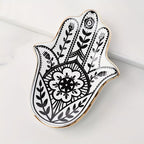 A Hamsa hand-shaped decorative tray with black floral and geometric henna-style patterns on a white background, finished with a polished gold rim, photographed on a light marble surface.