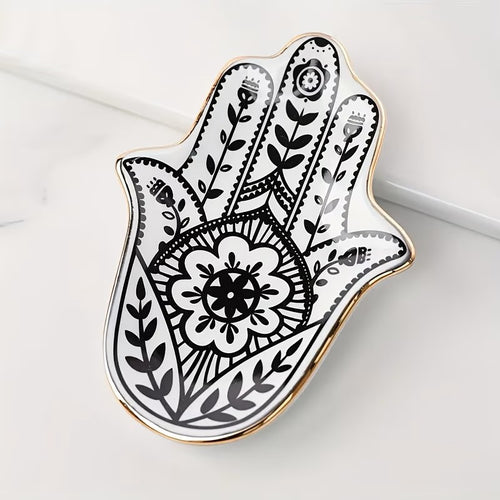 A Hamsa hand-shaped decorative tray with black floral and geometric henna-style patterns on a white background, finished with a polished gold rim, photographed on a light marble surface.