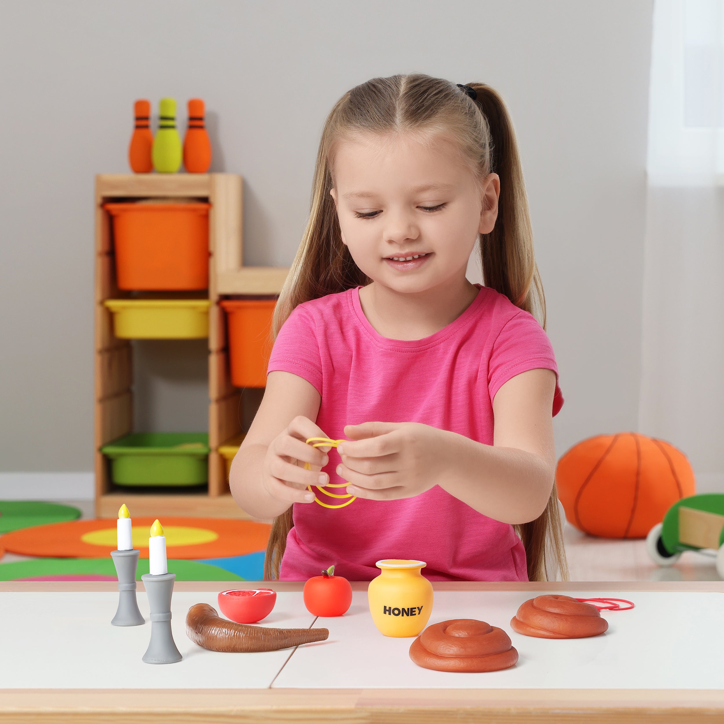 A young girl with blonde hair in a ponytail is sitting at a table playing with a wooden pretend-play food toy set. She is wearing a pink t-shirt and holding a yellow toy honey pot.