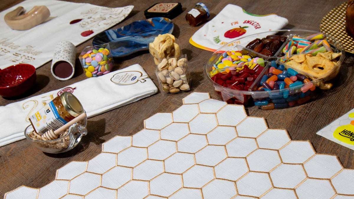 A styled tabletop arrangement featuring a white hexagonal tile mat as the central product, surrounded by assorted snacks, candies, jars, and folded white towels on a wooden surface.
