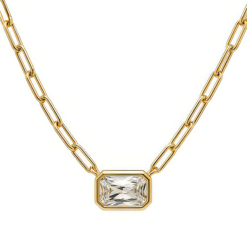 A gold paperclip chain necklace with a rectangular bezel-set clear gemstone pendant, shown against a plain white background.