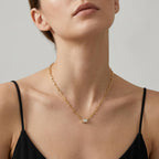 A close-up of a model wearing a delicate gold chain necklace with a small rectangular clear gemstone pendant, styled with a black spaghetti-strap top against a neutral gray background.