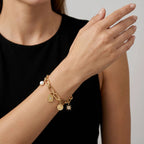 A close-up of a model's wrist wearing a gold charm bracelet with a white pearl bead and multiple small gold charms, including a shield, key, and star.