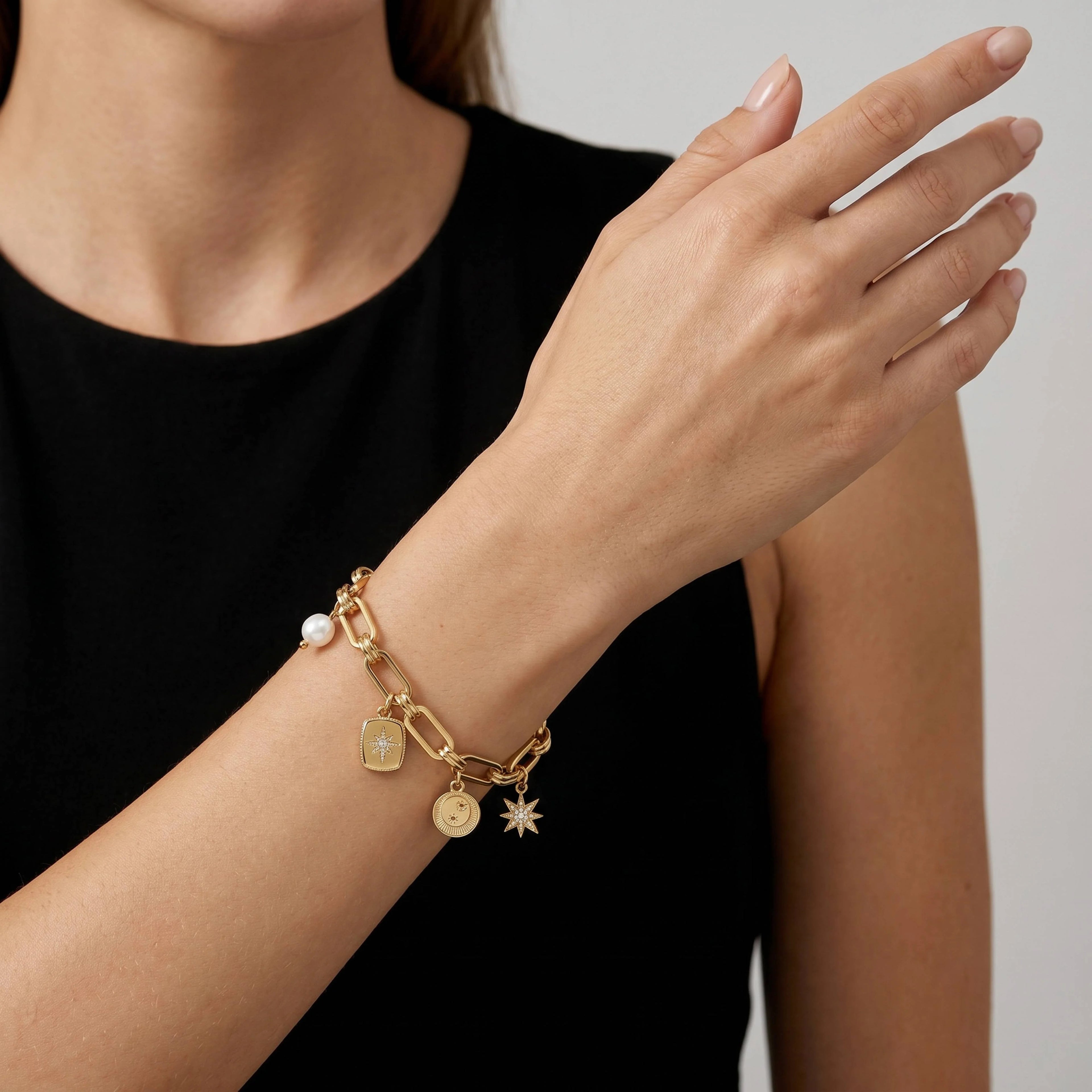 A close-up of a model's wrist wearing a gold charm bracelet with a white pearl bead and multiple small gold charms, including a shield, key, and star.