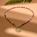 A beaded necklace with a gold toggle clasp and a circular charm featuring a colorful evil eye design, displayed on a warm beige background with soft shadows and a green fern leaf at the top.
