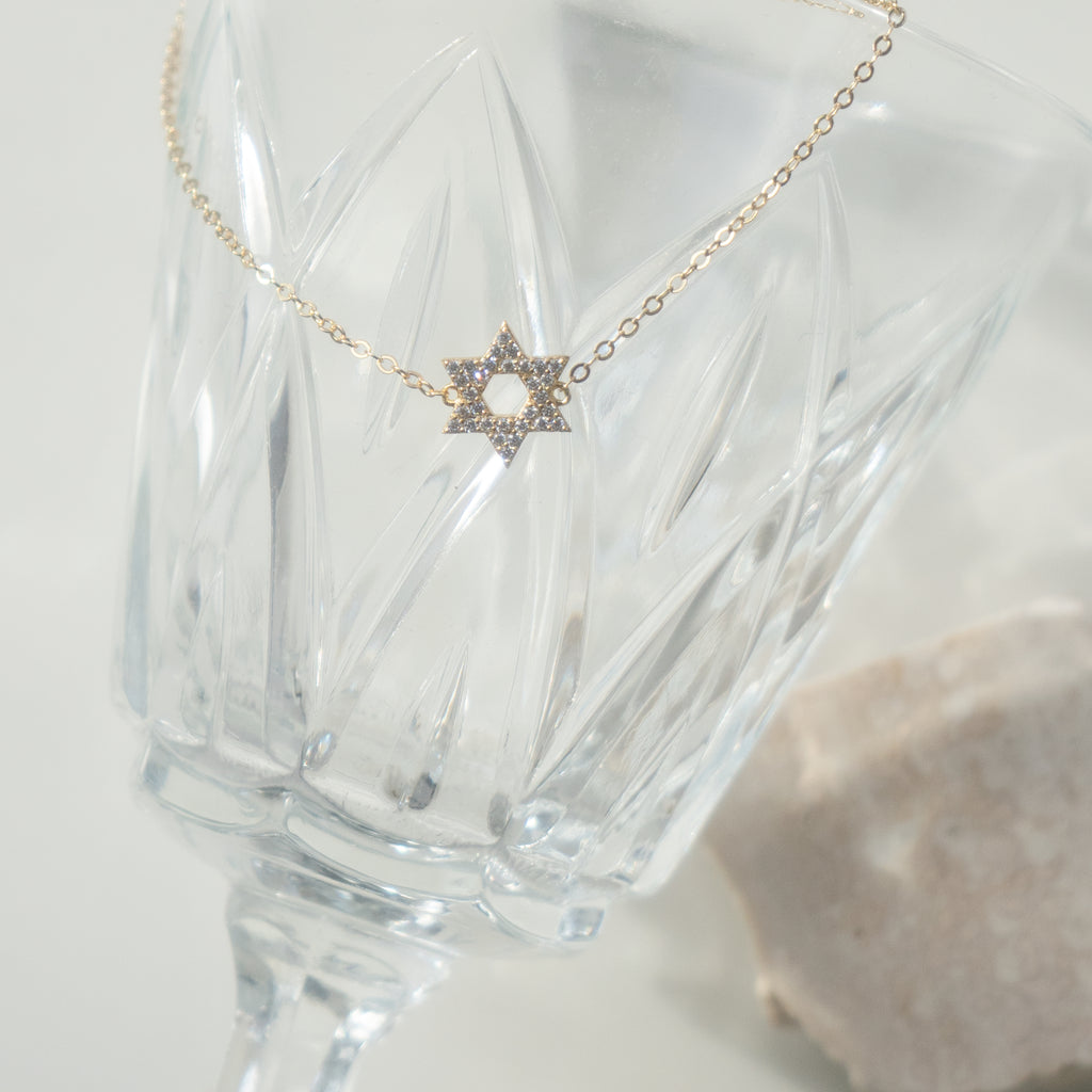 A delicate gold chain necklace with a Star of David pendant adorned with small clear stones, displayed draped over a faceted clear glass on a soft light background.