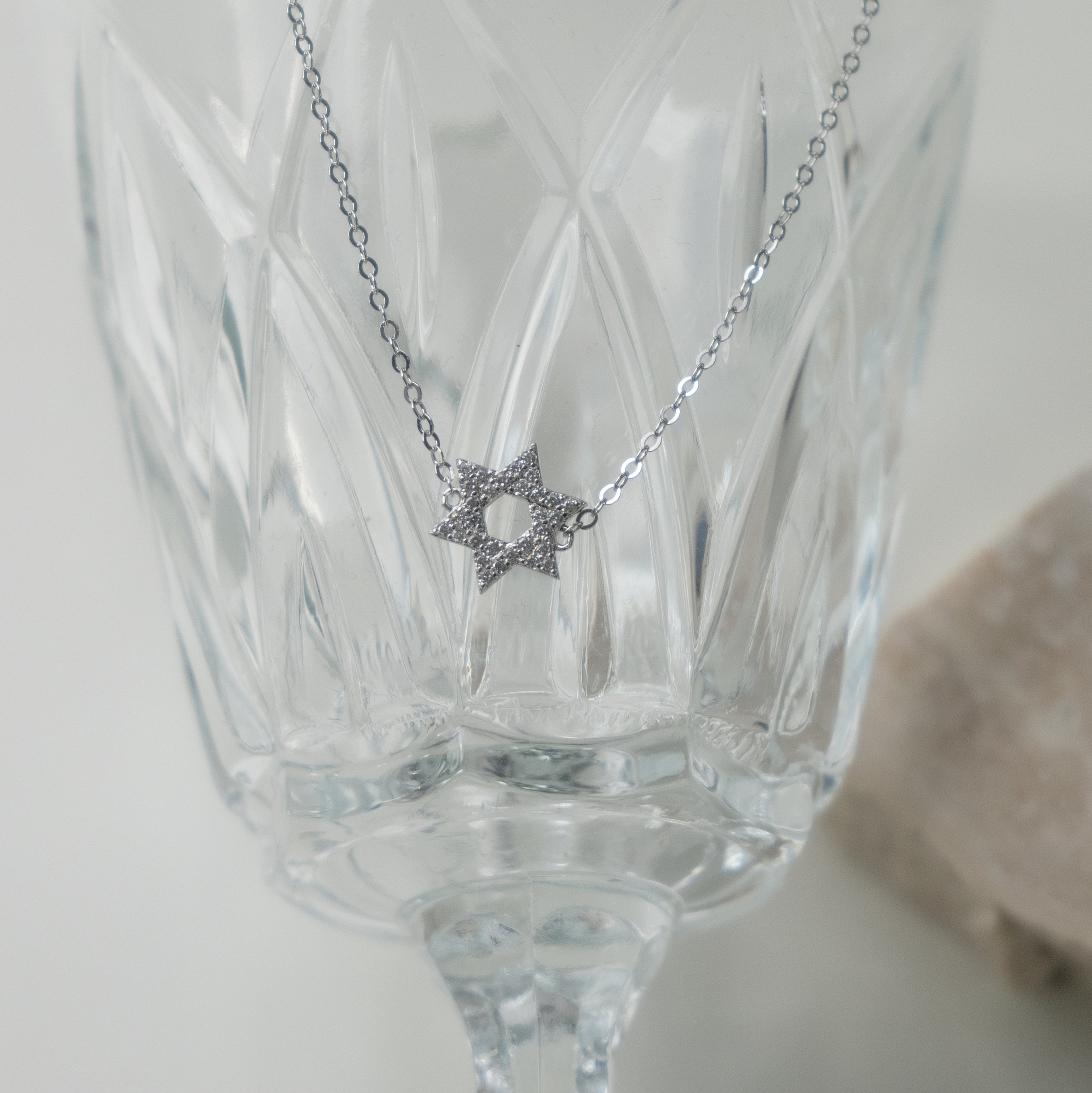 A silver pendant with the Star of David charm, hanging in front of a glass goblet.