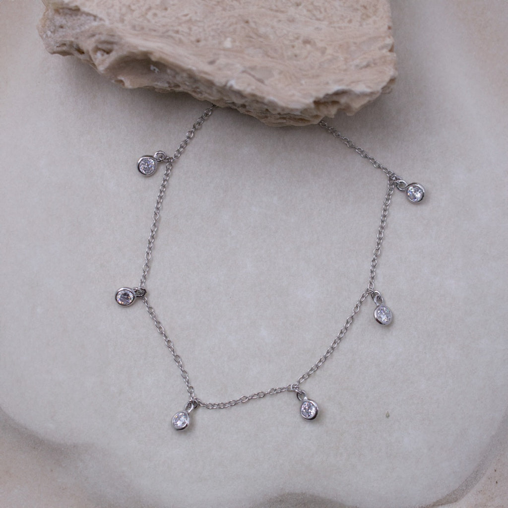 A delicate silver chain necklace with five small teardrop-shaped clear gemstones, displayed on a textured beige stone surface.
