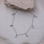 A delicate silver chain necklace with five small teardrop-shaped clear gemstones, displayed on a textured beige stone surface.
