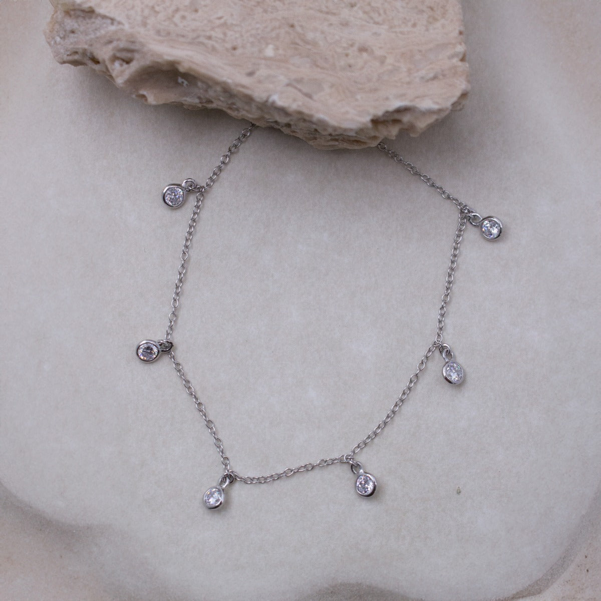 A delicate silver chain necklace with five small teardrop-shaped clear gemstones, displayed on a textured beige stone surface.