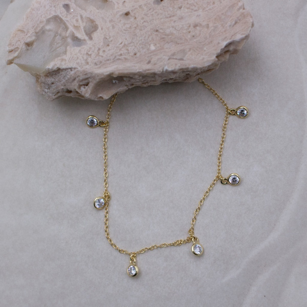 A delicate gold chain necklace with five small clear teardrop-shaped crystals, displayed on a light beige textured surface with a decorative stone prop.