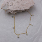 A delicate gold chain necklace with five small clear teardrop-shaped crystals, displayed on a light beige textured surface with a decorative stone prop.