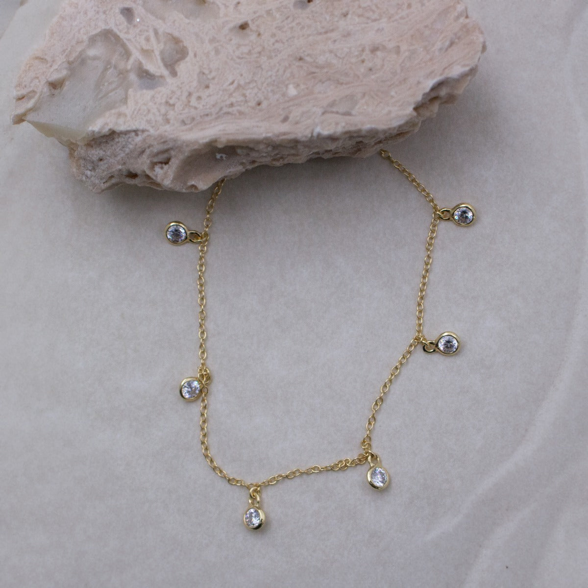 A delicate gold chain necklace with five small clear teardrop-shaped crystals, displayed on a light beige textured surface with a decorative stone prop.
