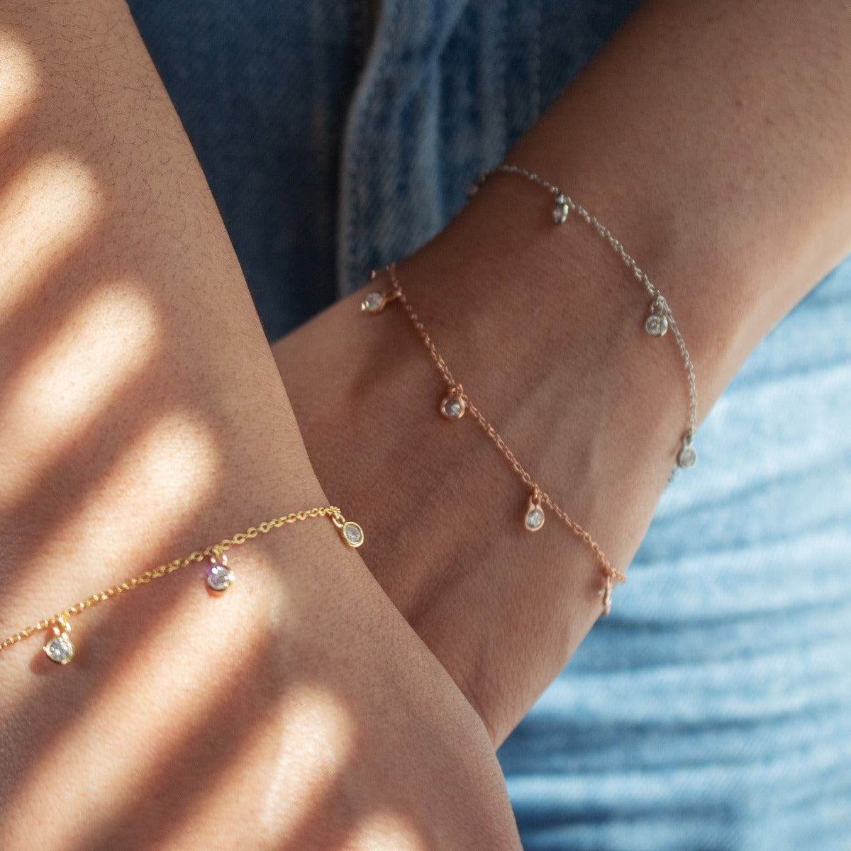 Three delicate chain bracelets with small teardrop crystal charms worn on a person's wrist and ankle, shown against blue denim clothing with dappled sunlight shadows.
