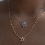 Two delicate Star of David necklaces worn on a model's neck. One necklace is silver with a silver chain and a silver pendant paved with small clear stones.