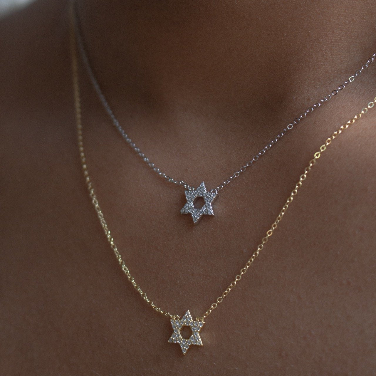 Two delicate Star of David necklaces worn on a model's neck. One necklace is silver with a silver chain and a silver pendant paved with small clear stones.