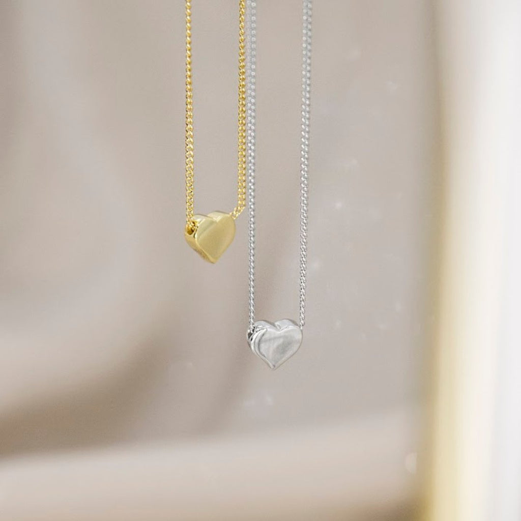 Two heart-shaped pendant necklaces hanging against a soft beige background, one in polished yellow gold and one in silver with a layered heart design.
