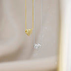 Two heart-shaped pendant necklaces hanging against a soft beige background, one in polished yellow gold and one in silver with a layered heart design.