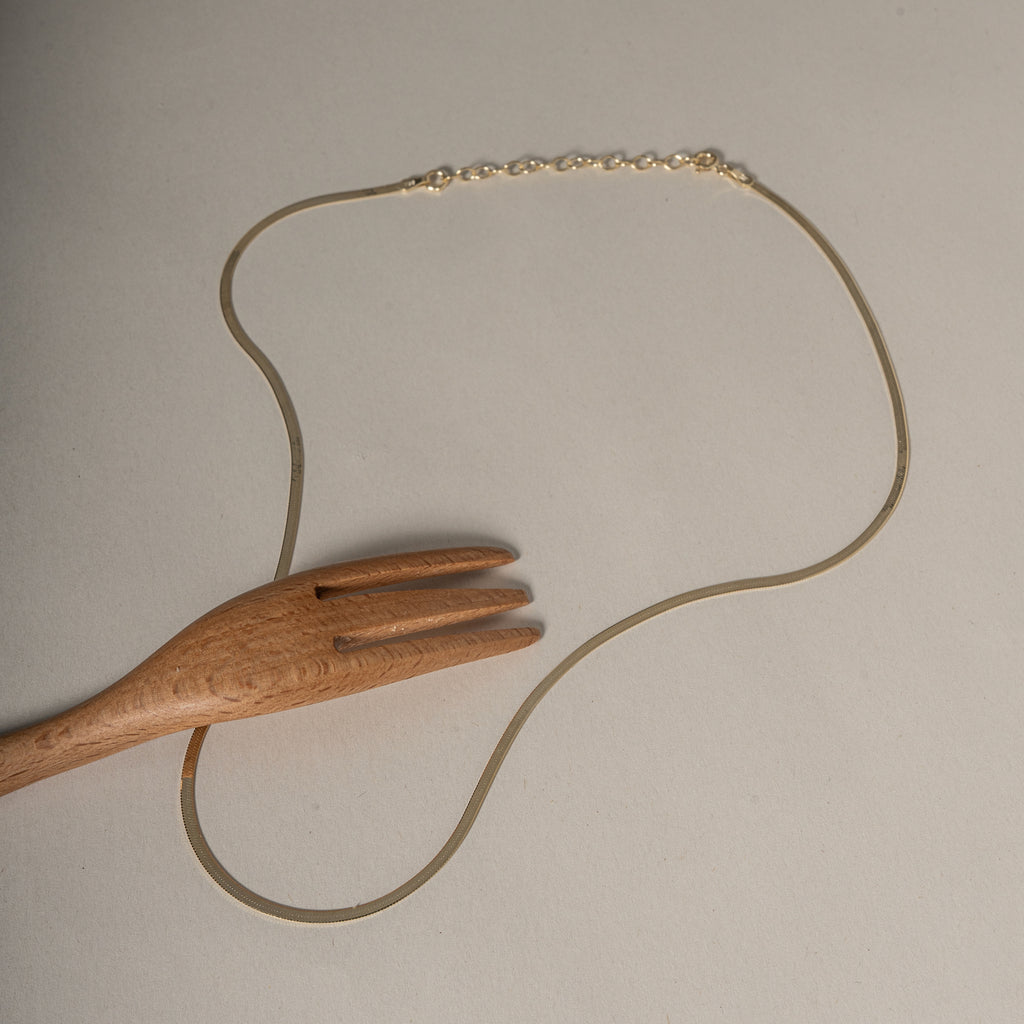 A delicate gold chain necklace with a lobster clasp, displayed on a neutral beige background with a wooden serving fork prop.