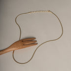 A delicate gold chain necklace with a lobster clasp, displayed on a neutral beige background with a wooden serving fork prop.