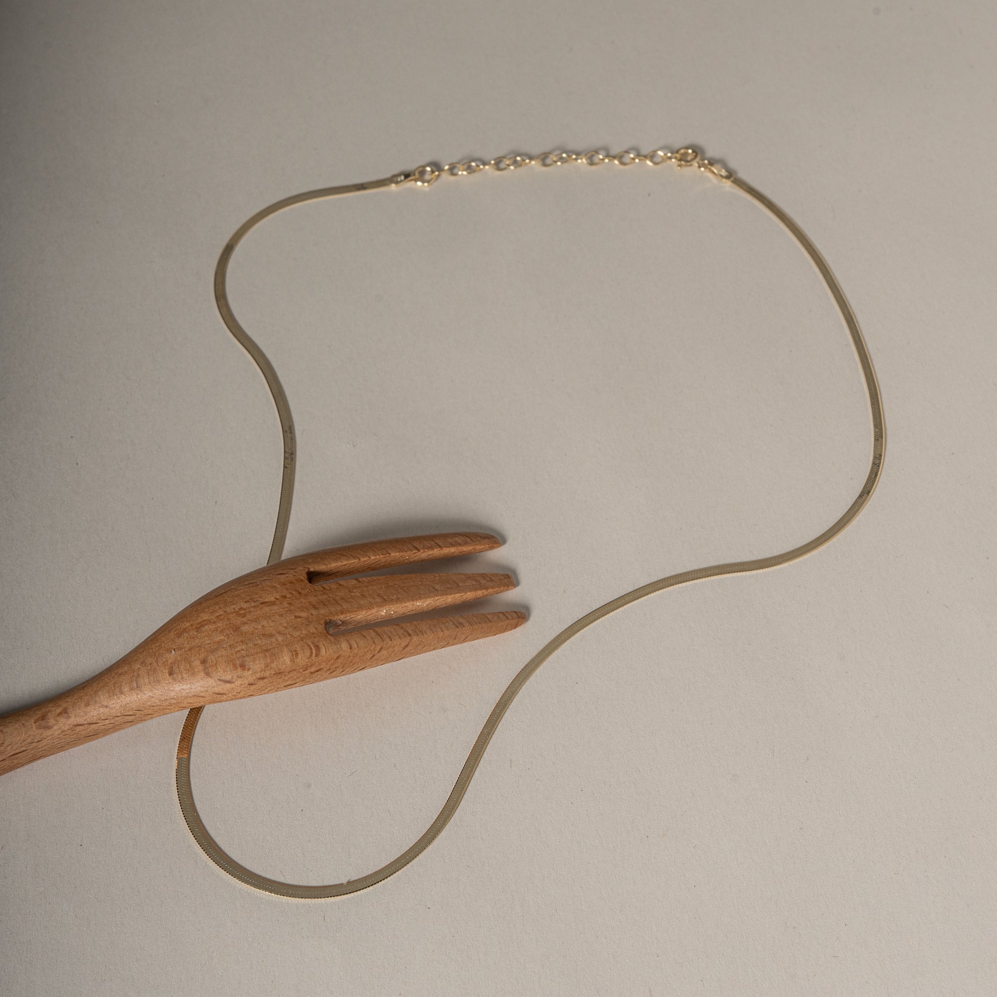 A delicate gold chain necklace with a lobster clasp, displayed on a neutral beige background with a wooden serving fork prop.