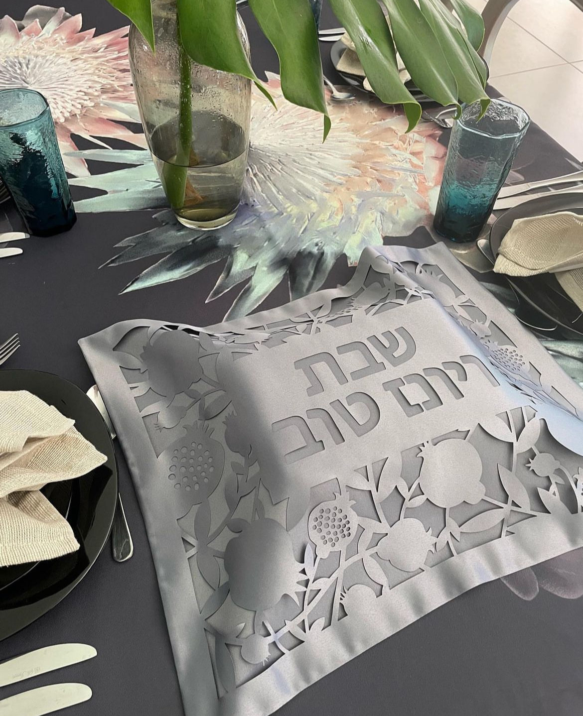 Decorative table setting with a silver cloth featuring Hebrew text, surrounded by flowers and glasses.