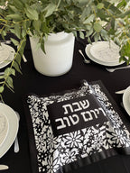 Table setting with black and white patterned napkin and Hebrew text on a black tablecloth.