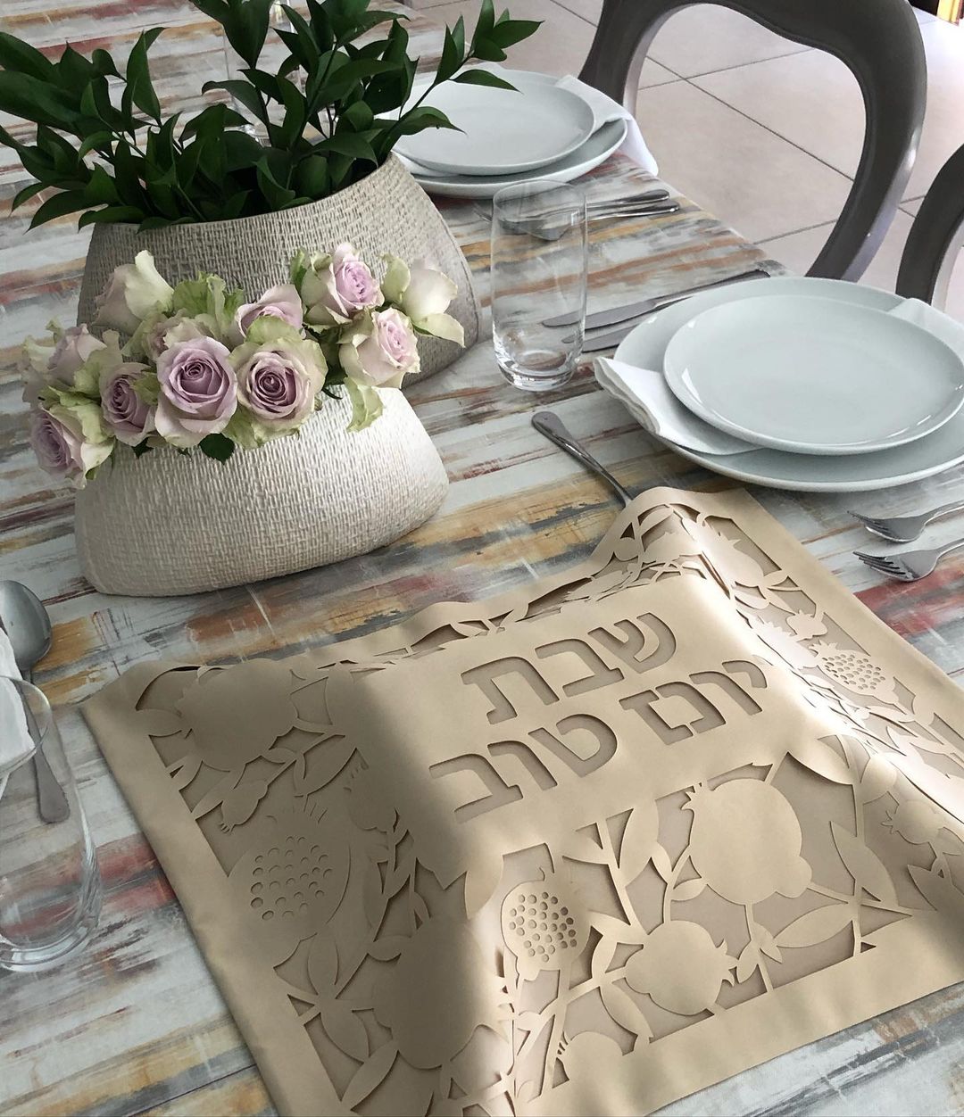 Dining table setting with floral arrangement, plates, glasses, and a decorative mat with Hebrew text.