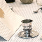 Silver kiddush cup on a saucer with Hebrew text on a textured tablecloth.