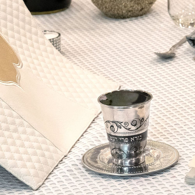 Silver kiddush cup on a saucer with Hebrew text on a textured tablecloth.